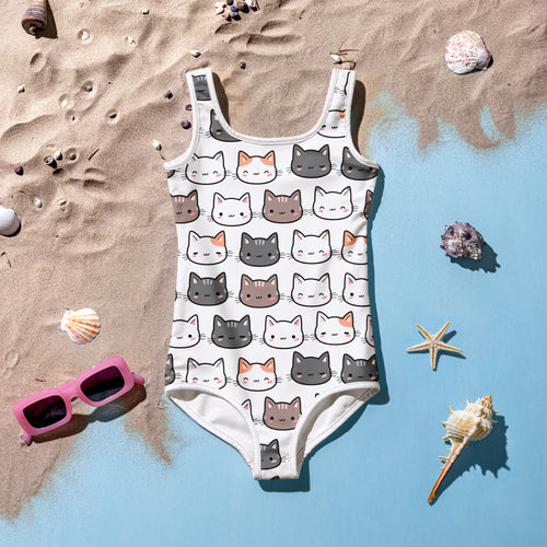 White kids swimsuit with cute cartoon cat faces, flat lay on sandy beach with shells and sunglasses