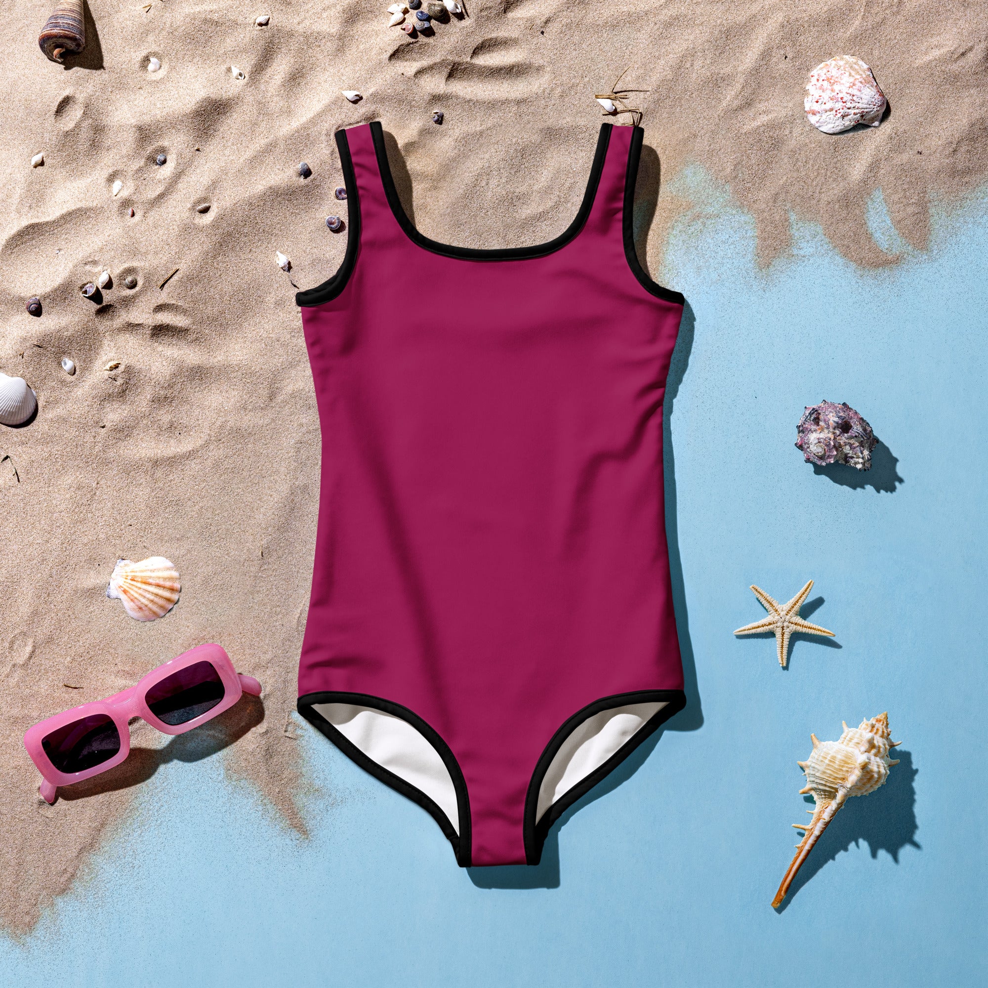 A vibrant magenta girls berry color buttery soft one-piece swimsuit with black trim white lining and upf 50 plus laid on sand