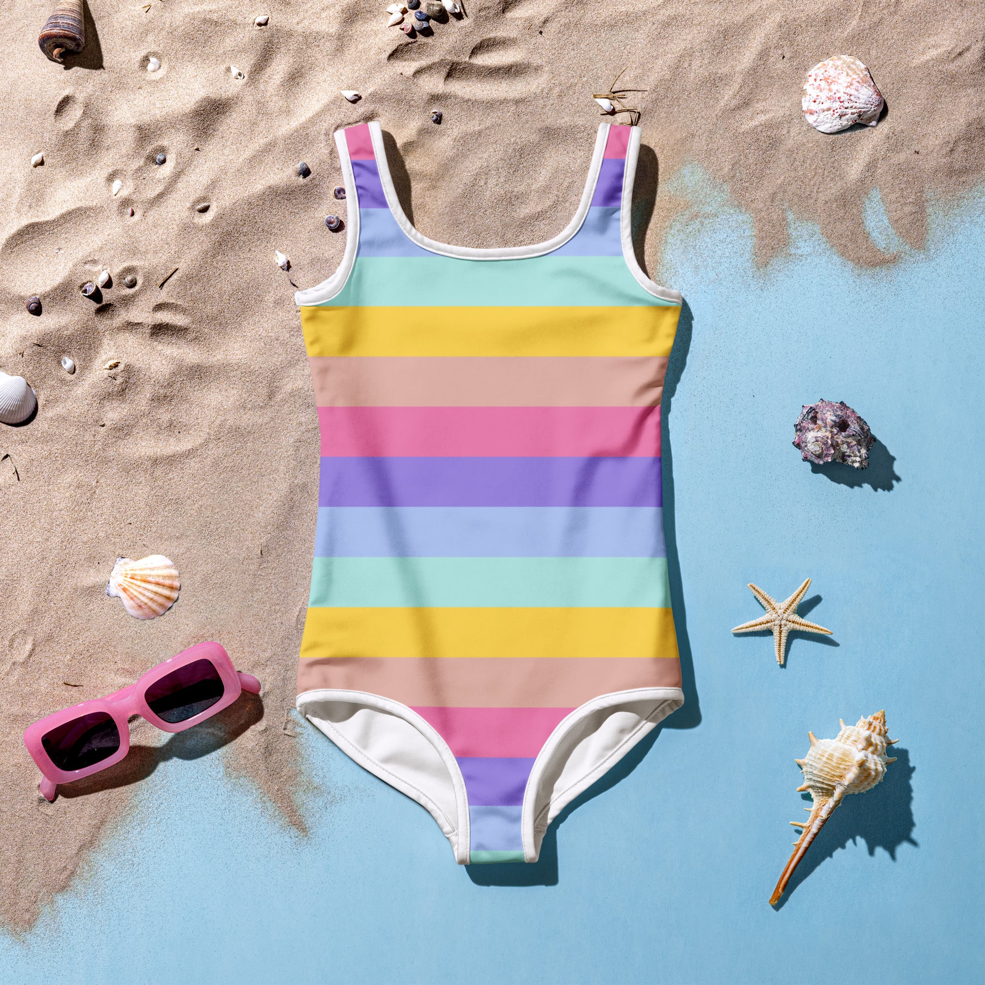 A vibrant rainbow-striped girls buttery soft one-piece swimsuit with white trim and UPF 50+ laid on sandy beach decor