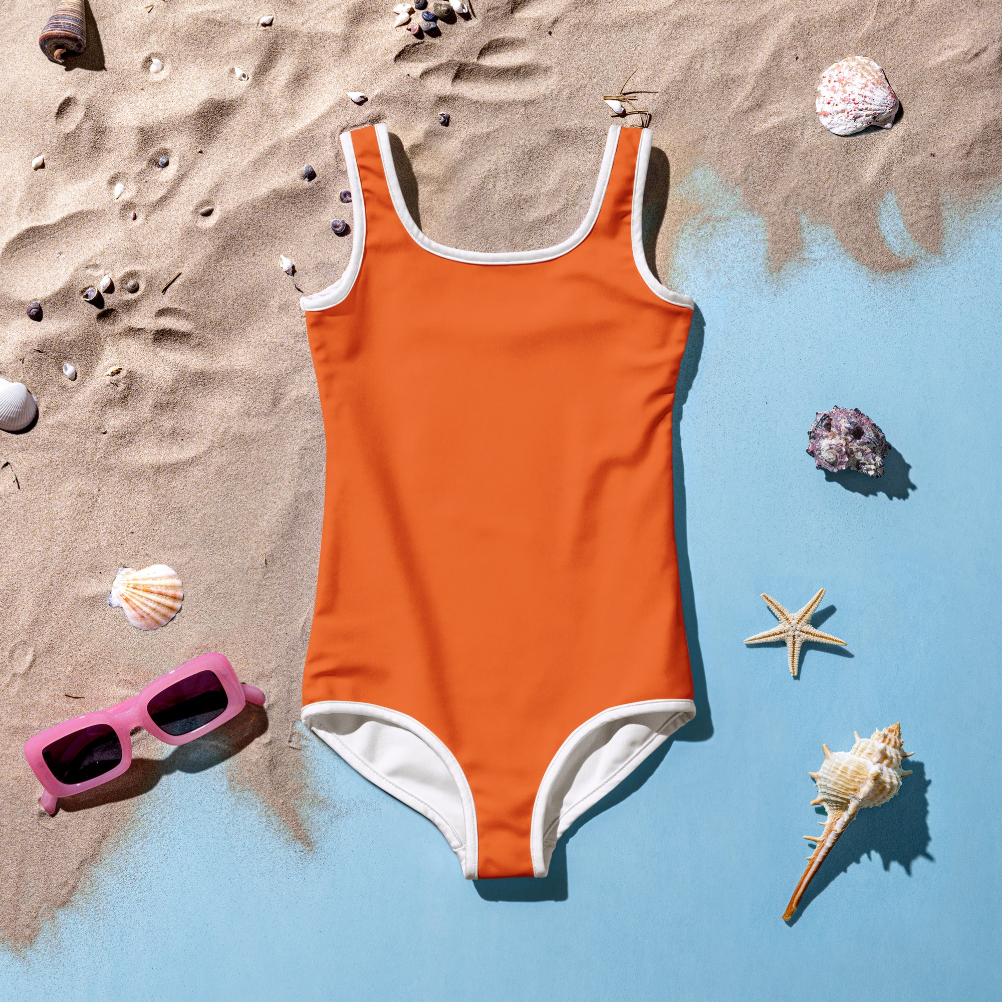 A vibrant orange girls buttery soft one-piece swimsuit with white trim and upf 50 plus laid flat on sand with seashells