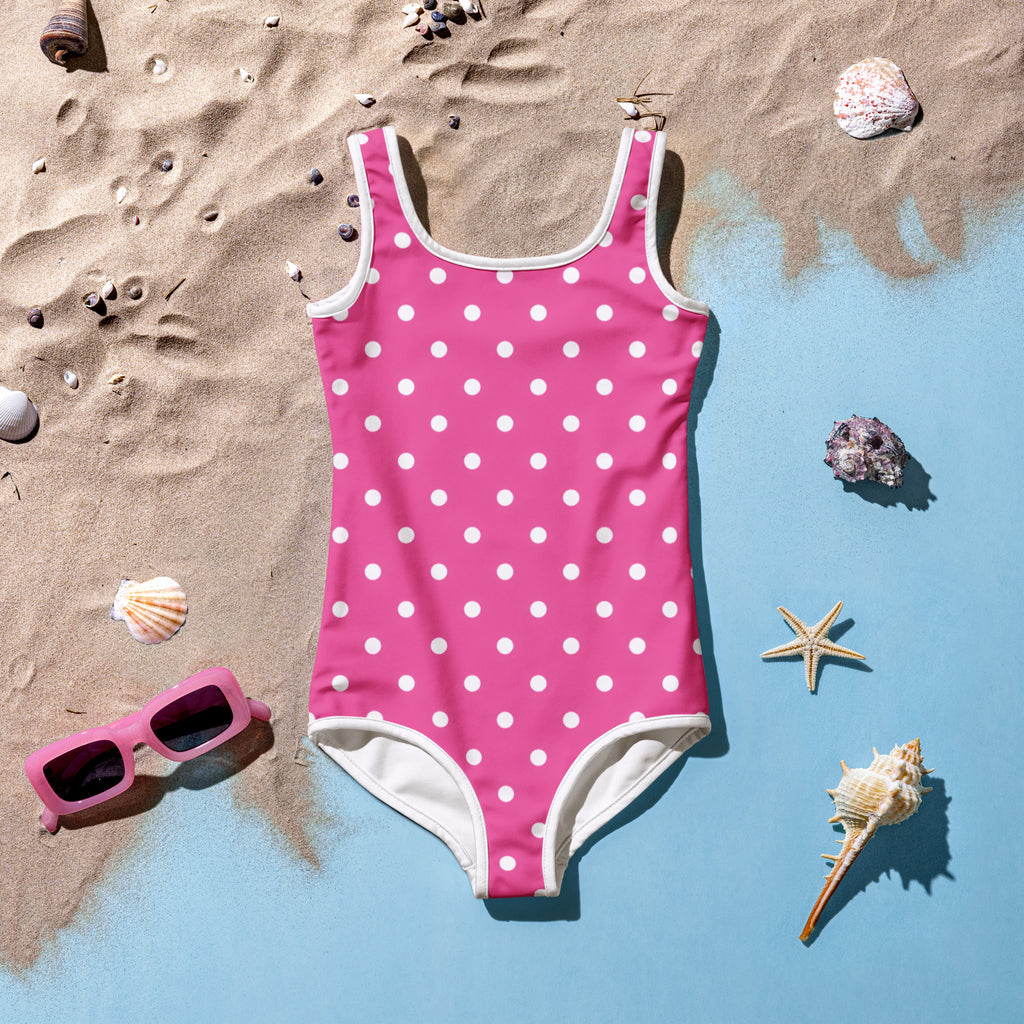 A pink one-piece girls swimsuit with white polka dots laid out on a sandy surface with beach shells and sunglasses around it, indicating a summer theme.