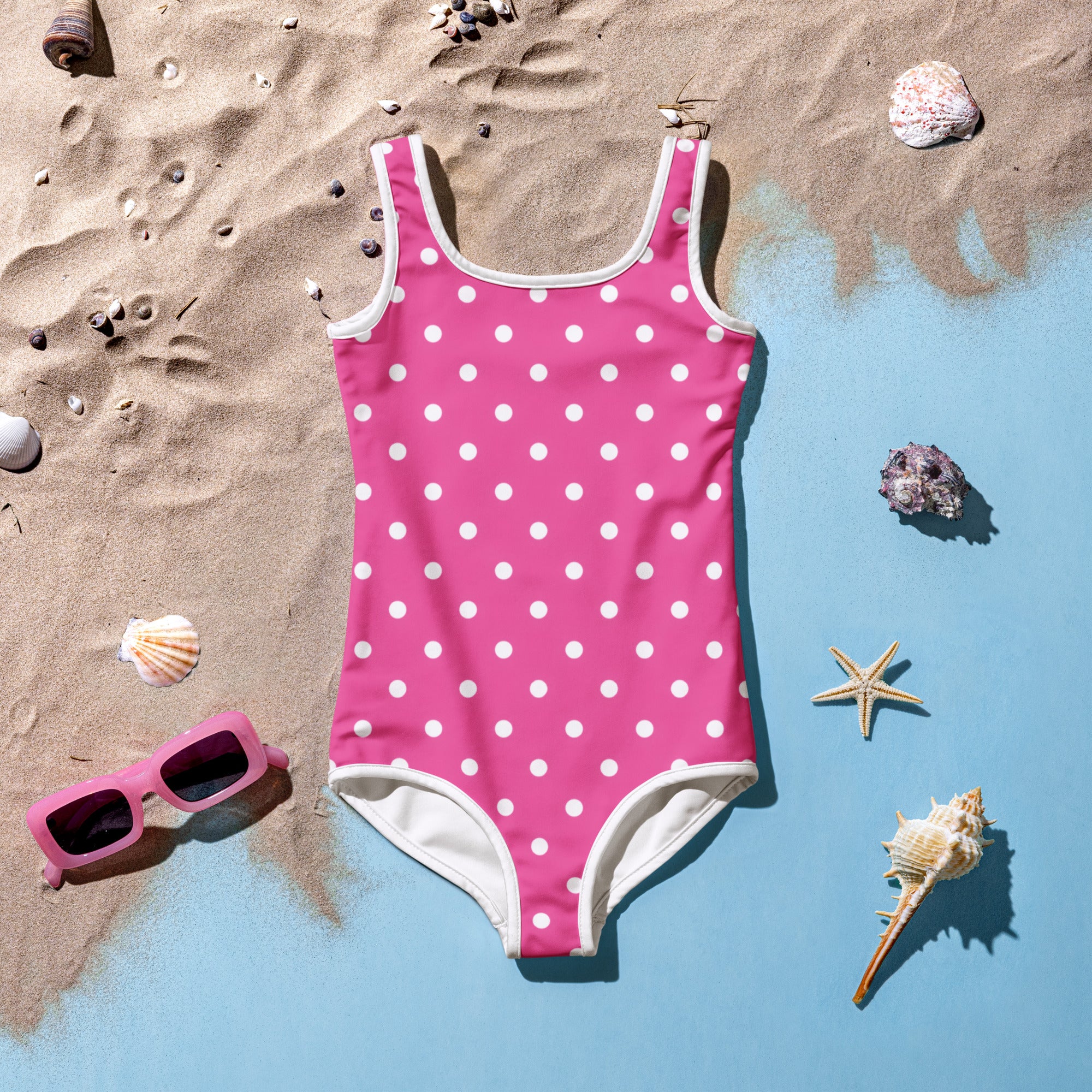 A pink one-piece girls swimsuit with white polka dots laid out on a sandy surface with beach shells and sunglasses around it, indicating a summer theme.