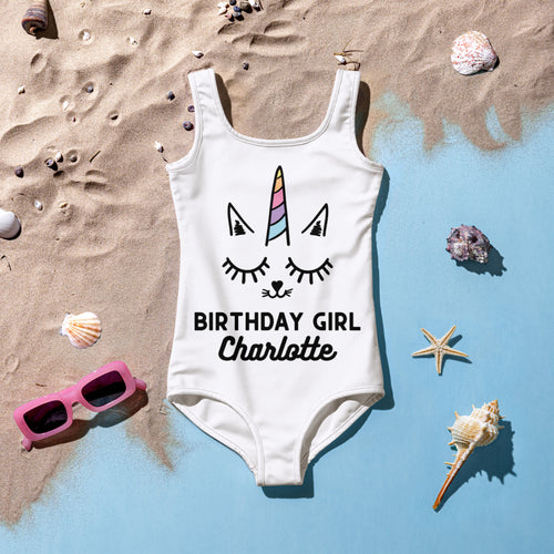 White one-piece swimsuit featuring a cartoon unicorn face with rainbow horn and birthday girl charlotte text in black