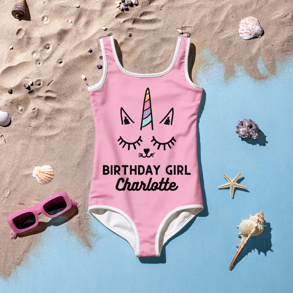 Pink one-piece swimsuit with white trim featuring a cartoon unicorn face and birthday girl charlotte text