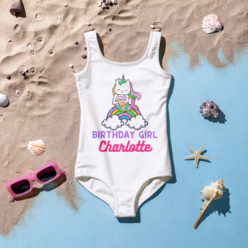 White swimsuit with unicorn design and 'Birthday Girl Charlotte' text on a sandy background with shells and sunglasses.