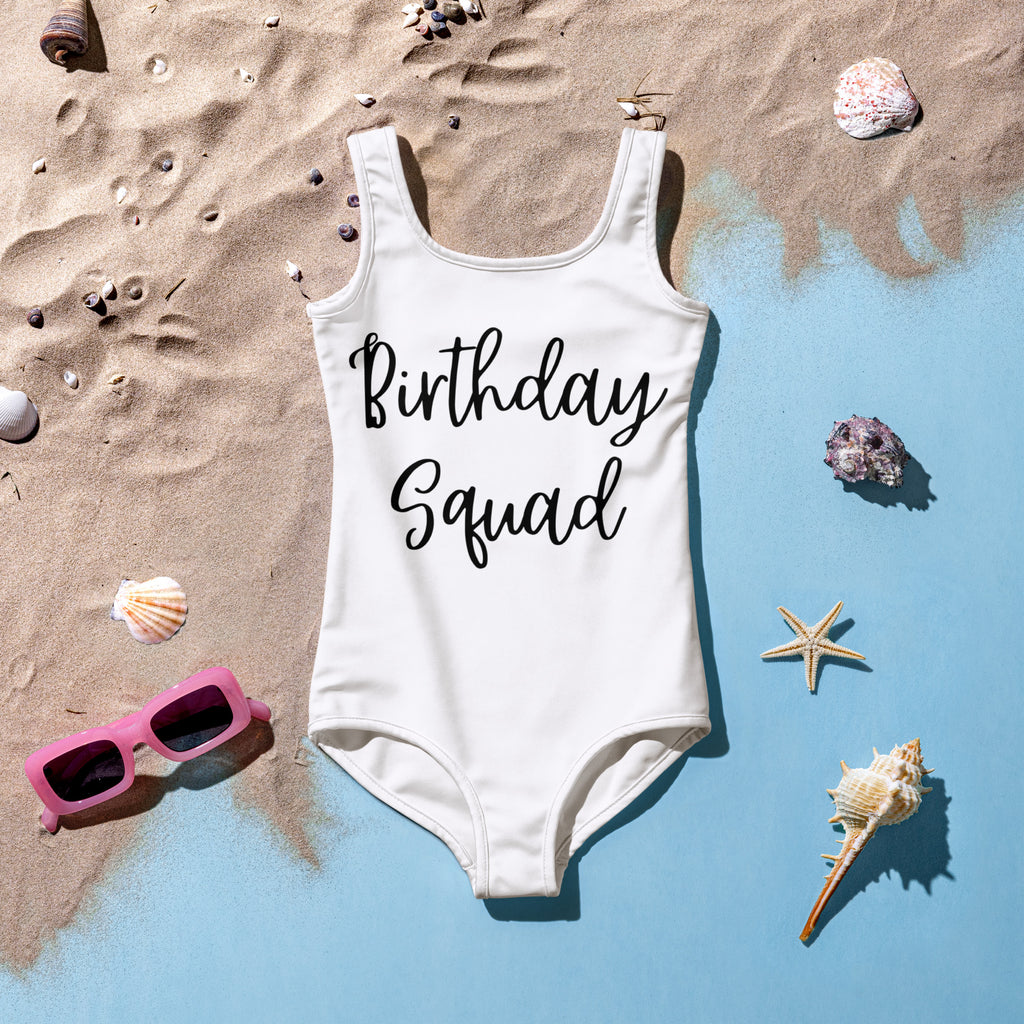 Girls white swimsuit with 'Birthday Squad' text on a sandy surface with shells and sunglasses.