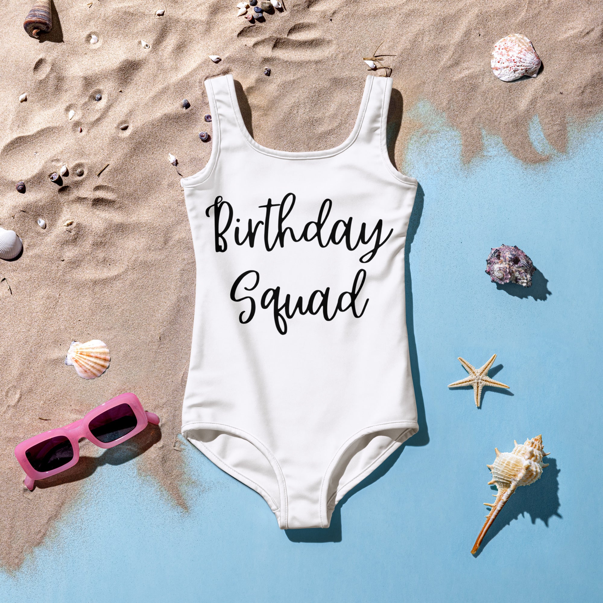 Girls white swimsuit with 'Birthday Squad' text on a sandy surface with shells and sunglasses.