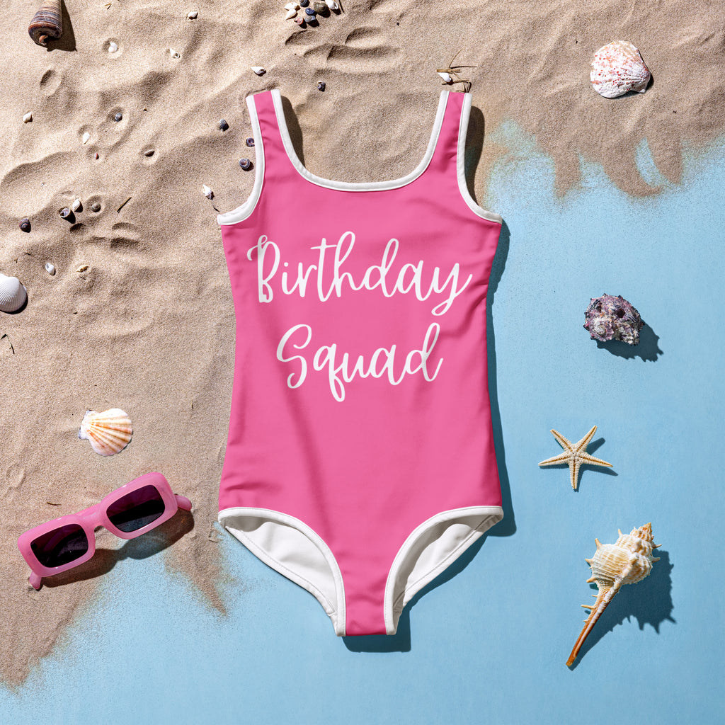 Kids pink swimsuit with 'Birthday Squad' text on a sandy background with shells and sunglasses.
