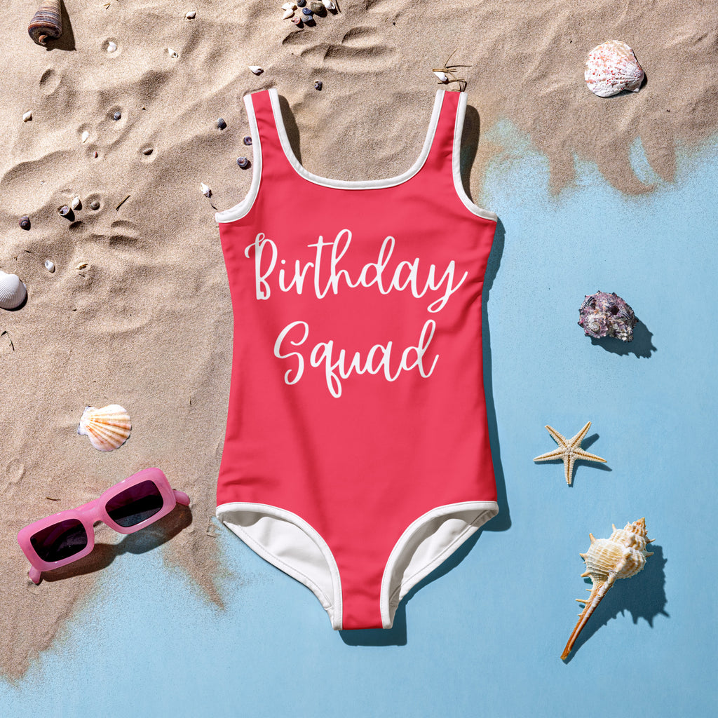 Kids red swimsuit with 'Birthday Squad' text on a sandy background with shells and sunglasses.
