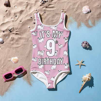 Girls pink swimsuit with 'It's My 9th Birthday' text on a sandy background with shells and sunglasses.