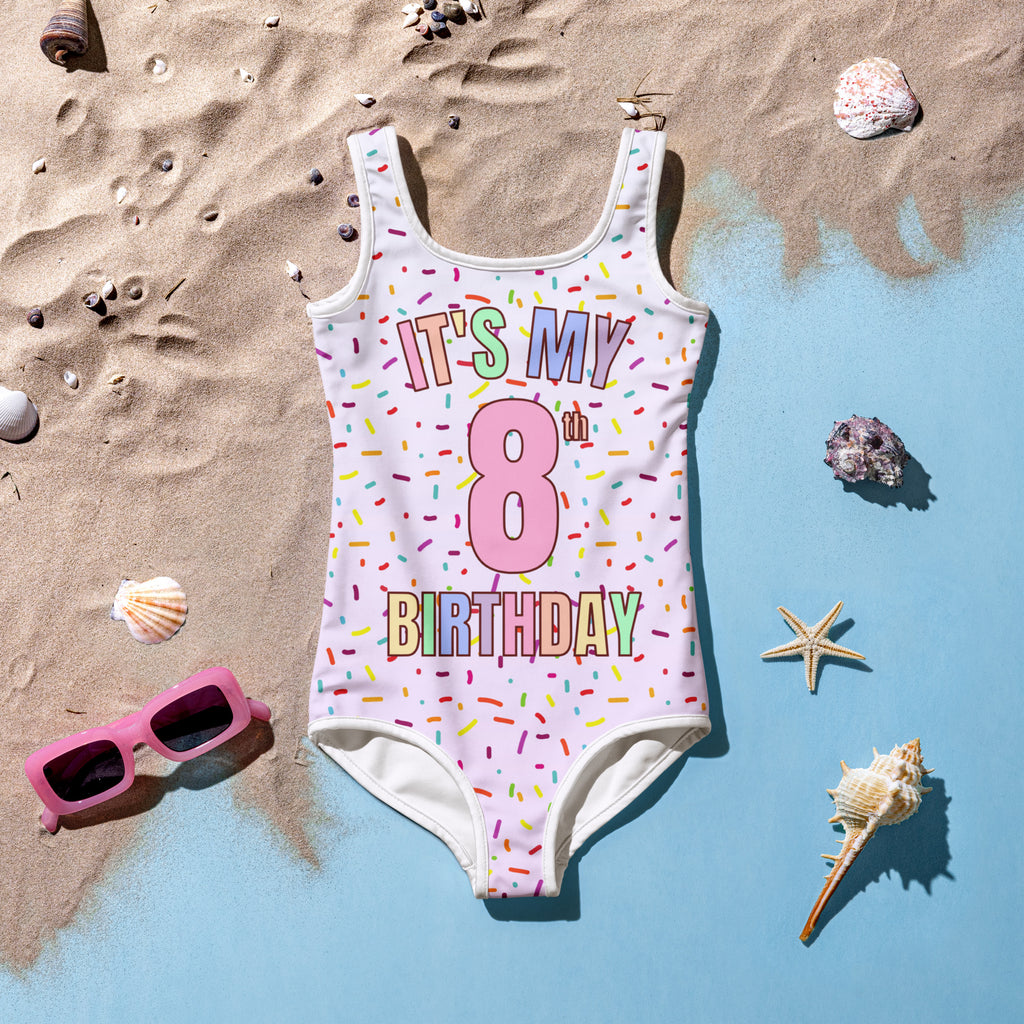 A white buttery soft girls one-piece swimsuit with rainbow sprinkles and its my 8th birthday text in rainbow letters