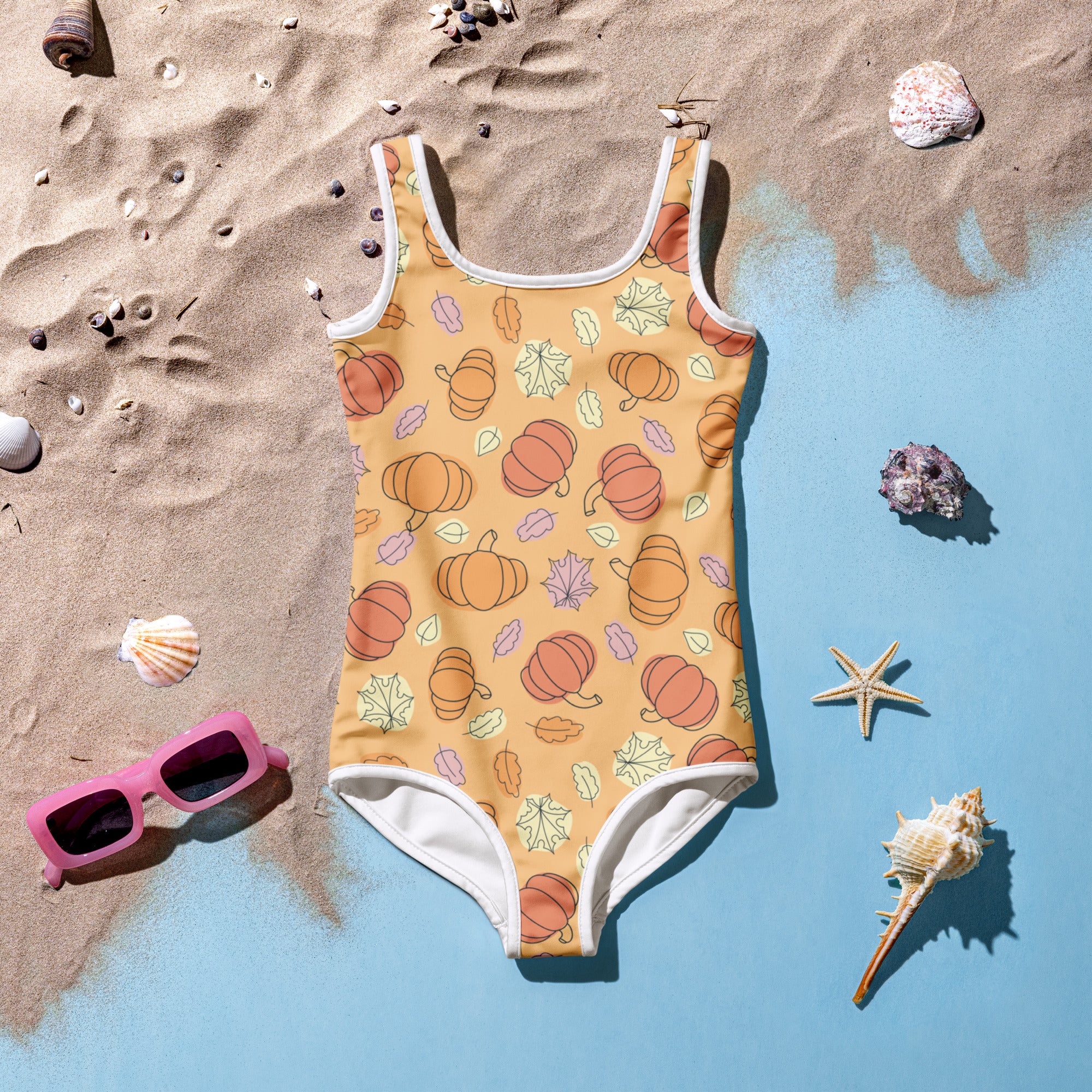 One-piece swimsuit with pumpkin pattern on a sandy surface with shells and sunglasses.