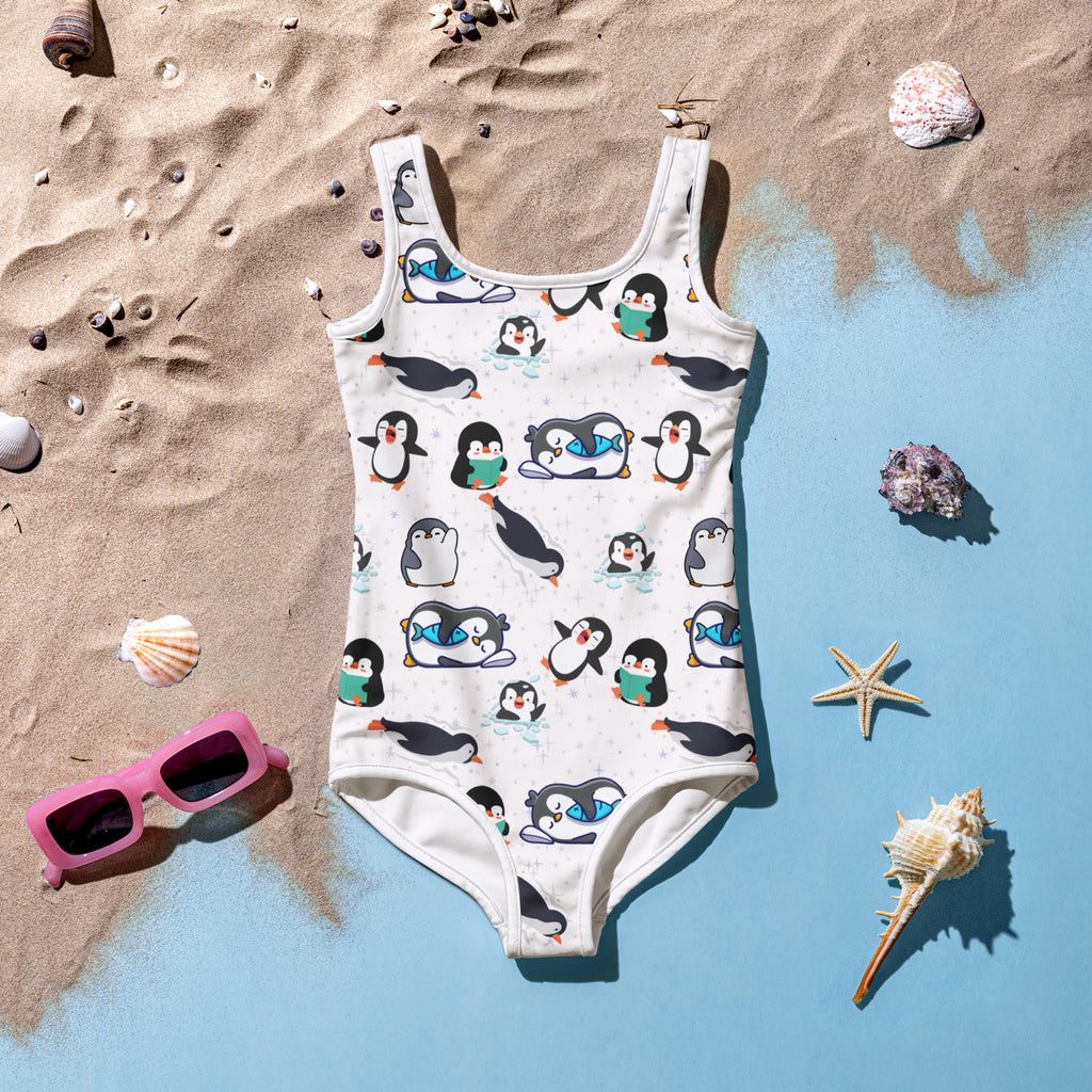 Children's swimsuit with penguin pattern on a sandy and blue gradient background with beach accessories.