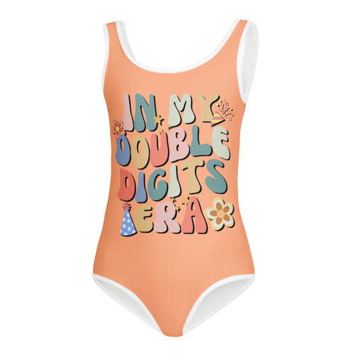 A peach-colored buttery soft girls one-piece swimsuit with white trim and colorful in my double digits era graphic