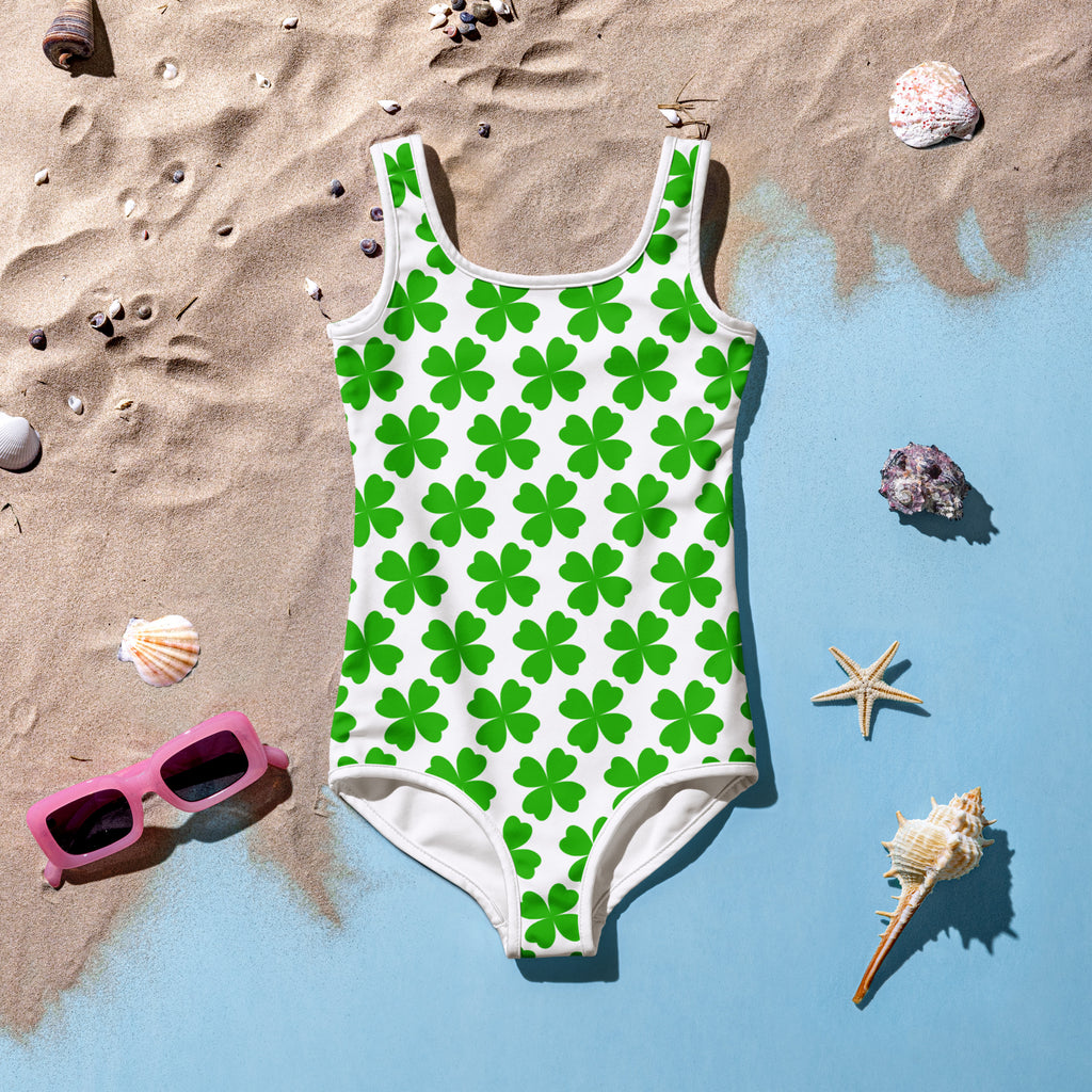A vibrant green shamrock-patterned girls one-piece swimsuit with white trim and buttery soft fabric laid on sand