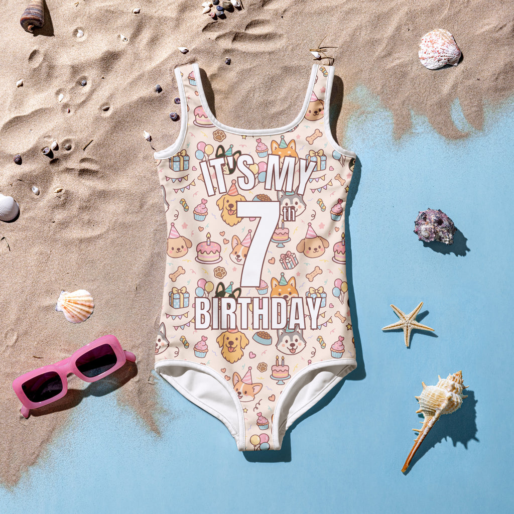Children's swimsuit with 'It's My 7th Birthday' text on a sandy background with shells and sunglasses.