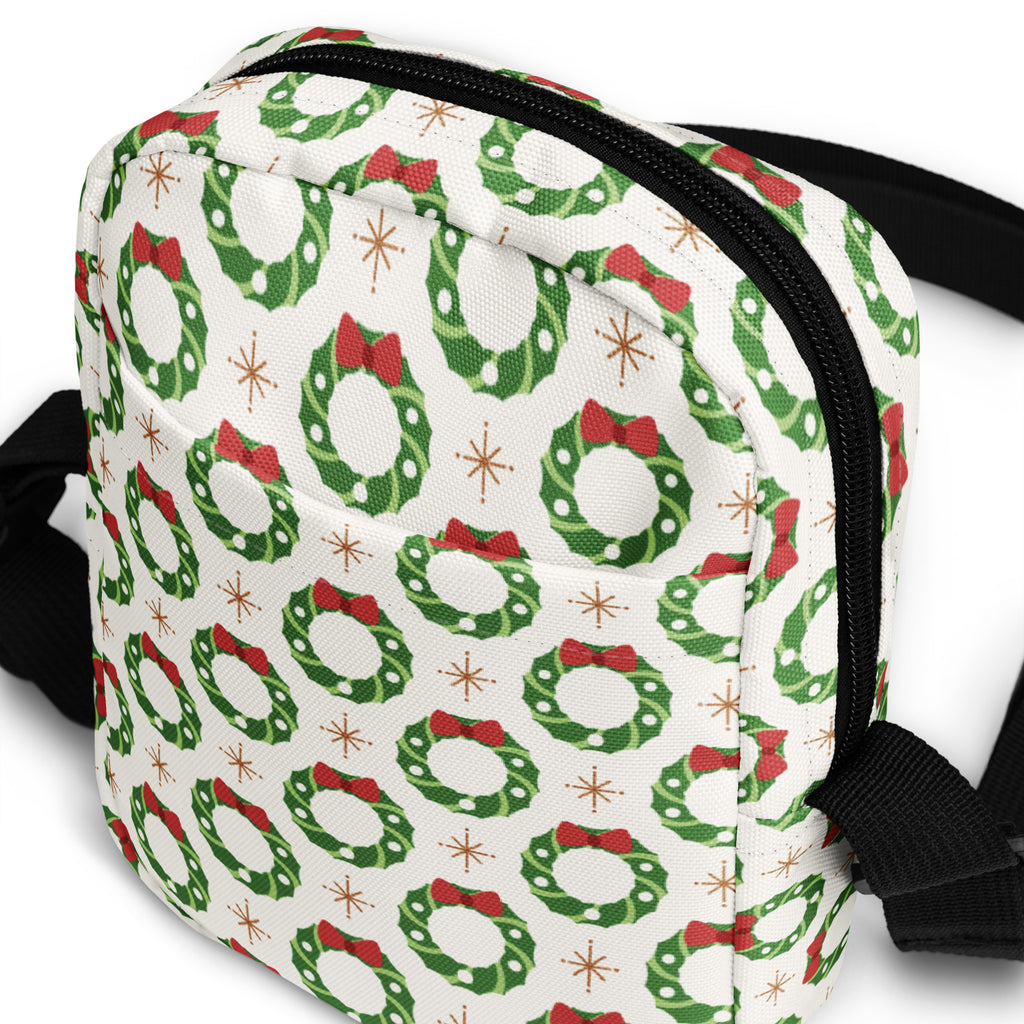 White crossbody bag with festive green wreaths red bows and gold starbursts on fabric surface for christmas use