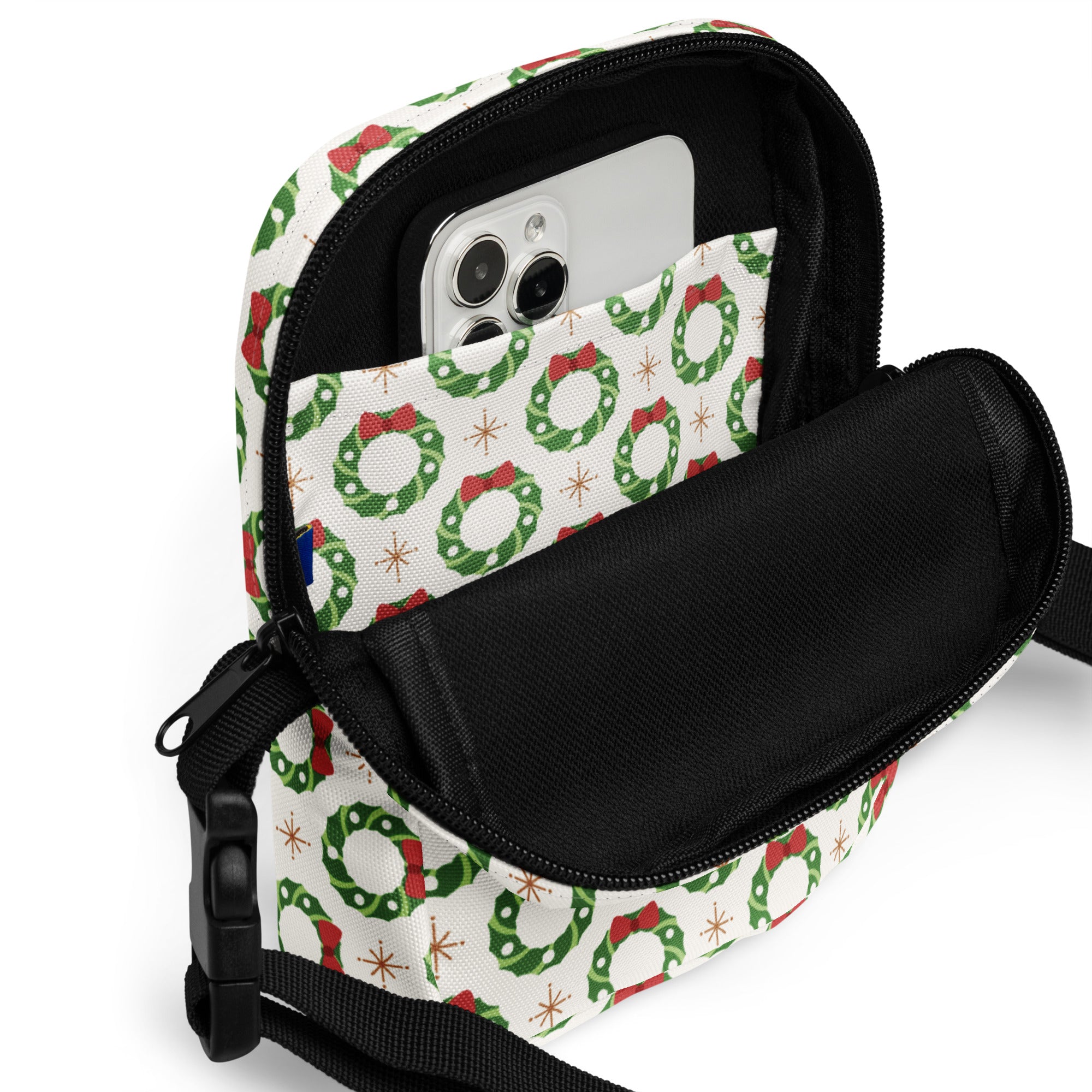 White crossbody purse with green wreath and red bow print on fabric featuring black zipper and adjustable strap