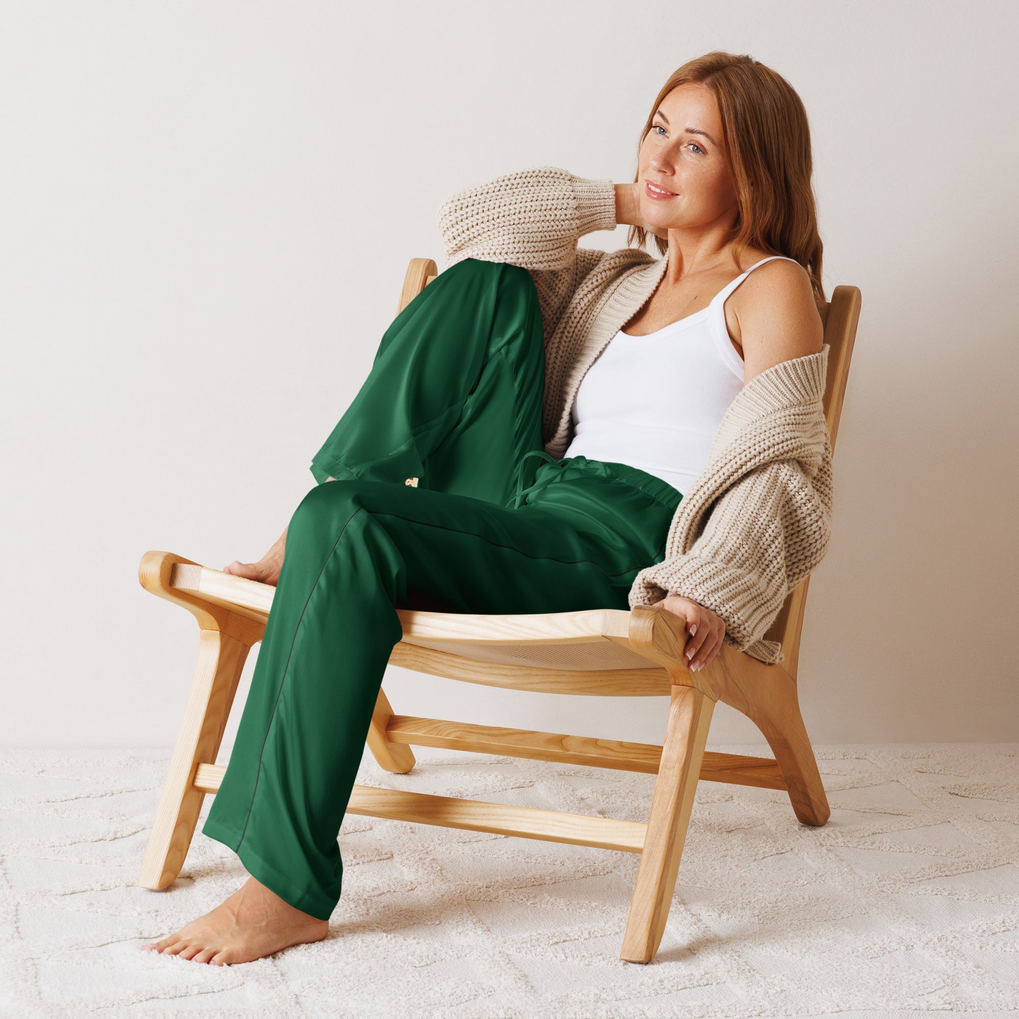 Woman wearing dark green satin pajama pants with drawstring waist seated in light wood chair with beige cardigan and white tank top