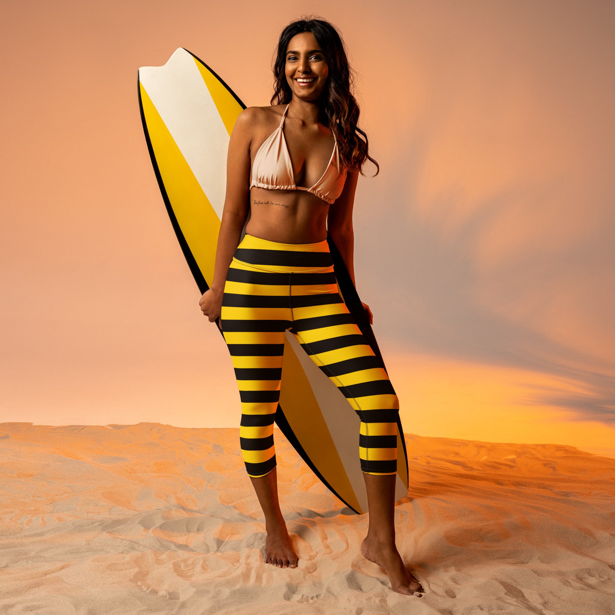 A woman holds a surfboard wearing high-rise buttery soft moisture-wicking yellow and black bumblebee stripe yoga capri leggings