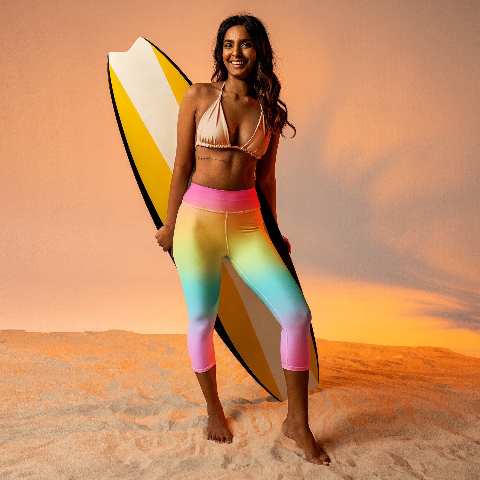 A woman wearing high-rise bright rainbow buttery soft yoga capri leggings and a beige bikini top stands barefoot on sand holding a striped surfboard
