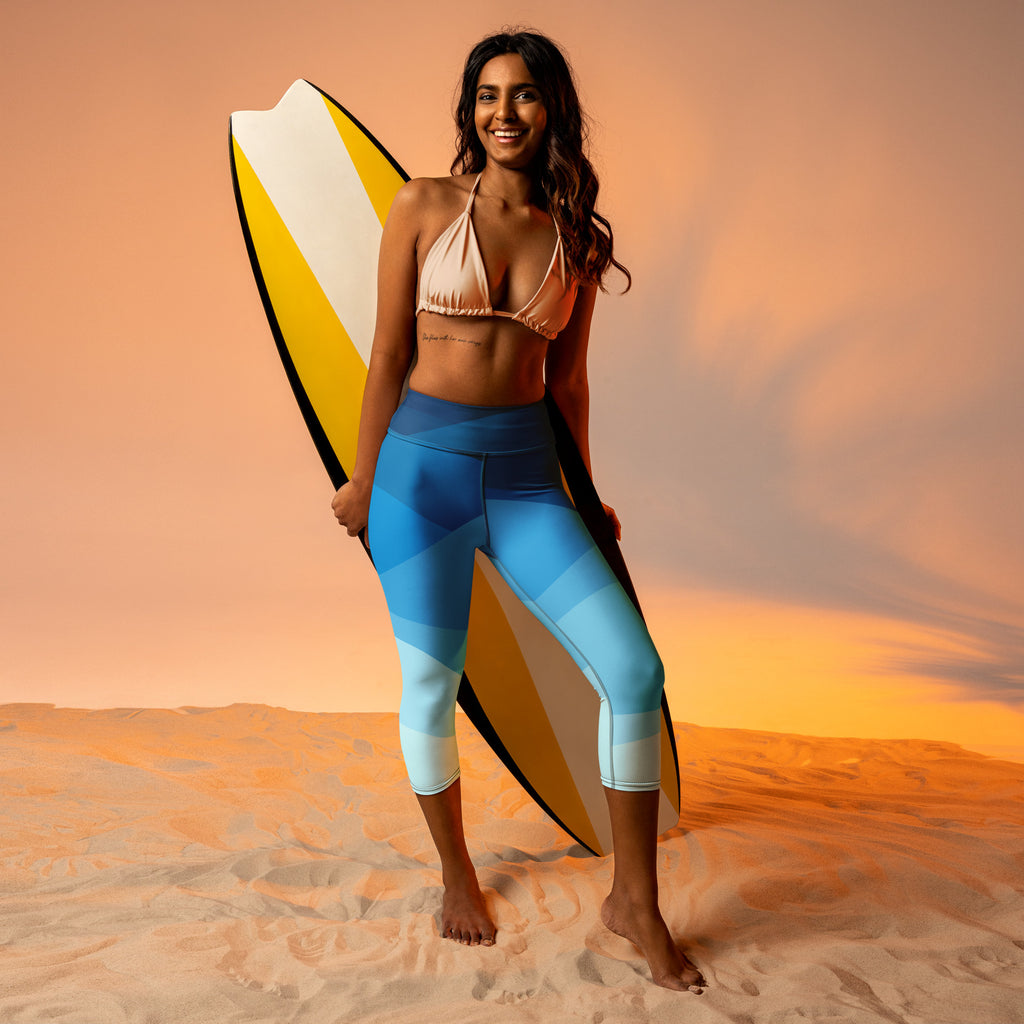 A woman stands barefoot on sand wearing wavy blue buttery soft high-rise moisture-wicking yoga capri leggings