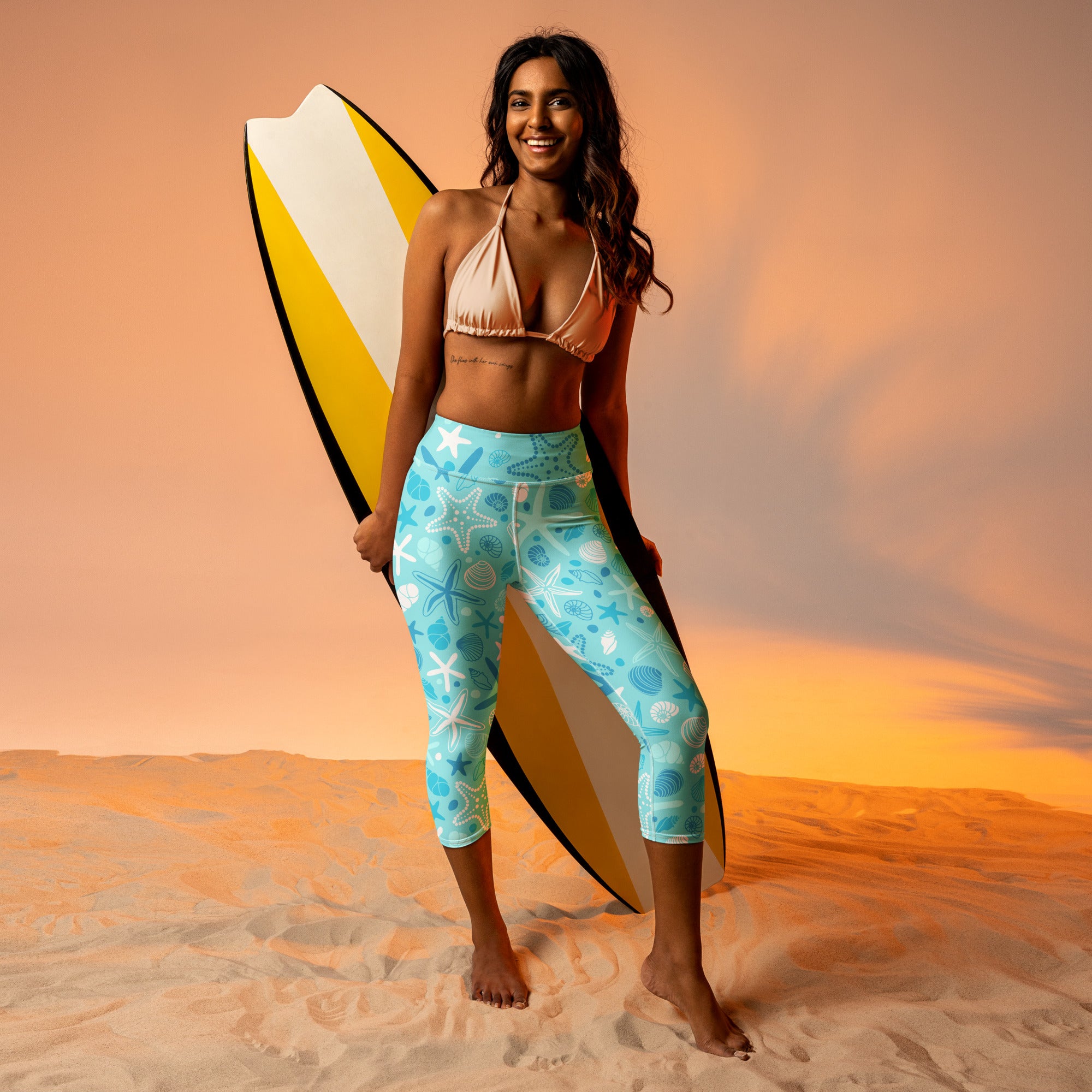 A woman wearing beige bikini top and turquoise high-rise buttery soft starfish and shells yoga capri leggings