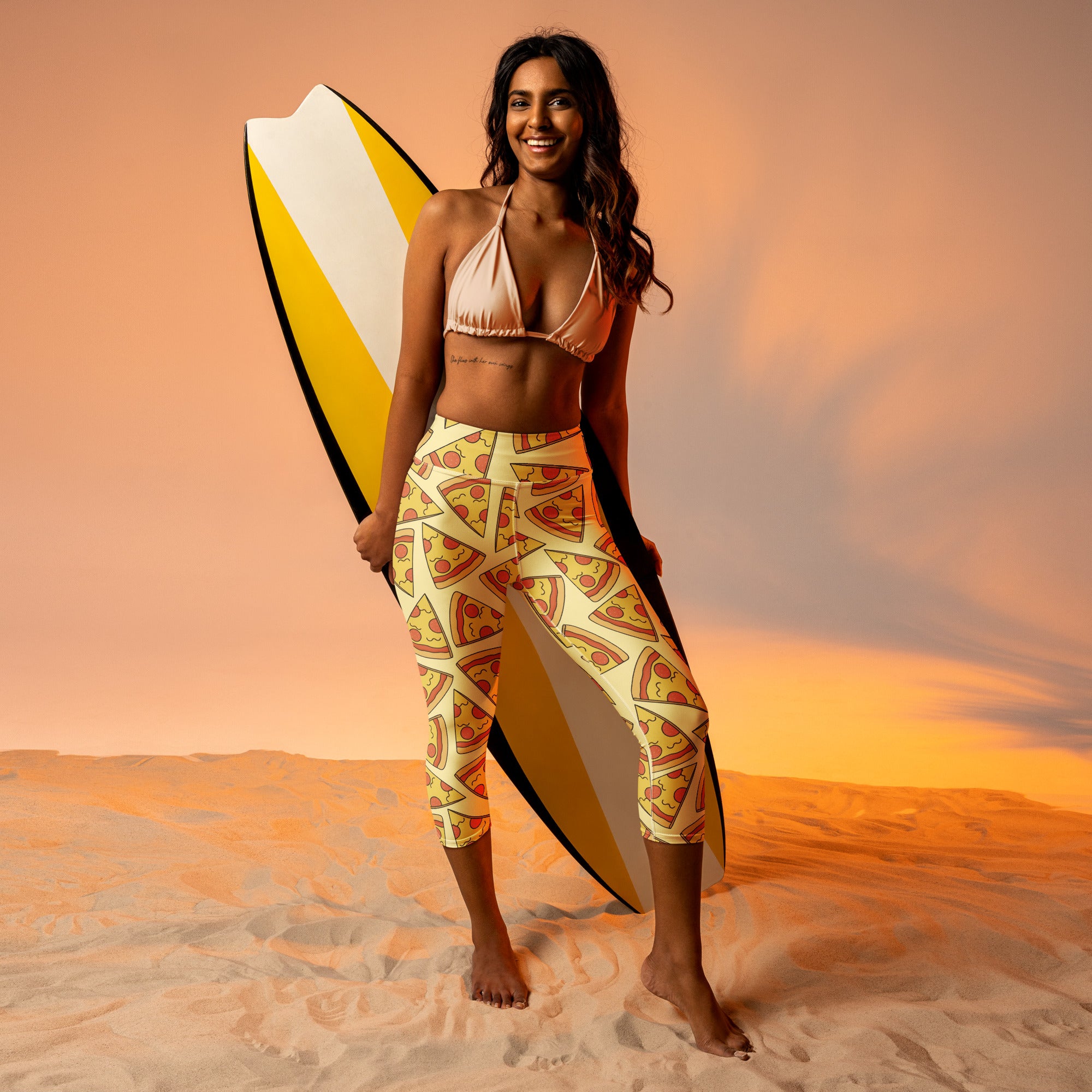 A woman stands barefoot in sand wearing beige bikini top and women’s pizza slice buttery soft yoga capri leggings