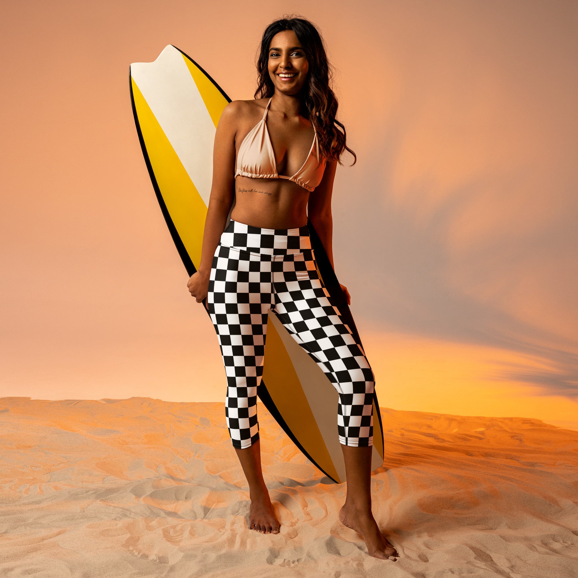 A woman wearing high-rise buttery soft black and white checkered yoga capri leggings holding a yellow surfboard