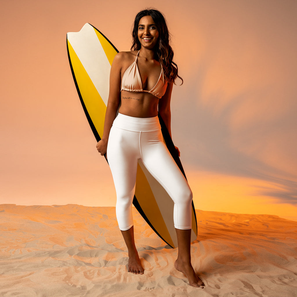 A woman wearing high-rise white buttery soft moisture-wicking yoga capri leggings stands barefoot on sand holding a surfboard