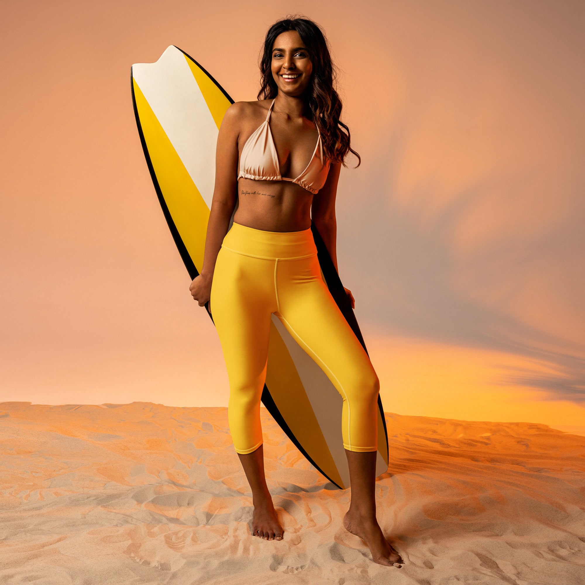 A smiling woman holds a vibrant yellow surfboard wearing yellow buttery soft high-rise moisture-wicking yoga capri leggings