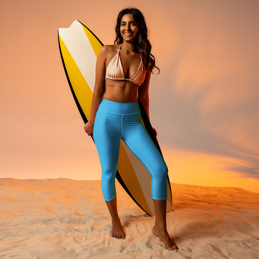A woman stands barefoot on sand wearing sky blue high-rise buttery soft moisture-wicking yoga capri leggings