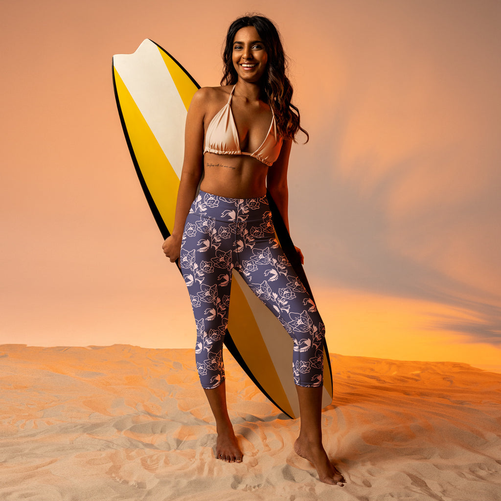 A woman wearing high-rise moisture-wicking navy blue floral yoga capri leggings holding a yellow surfboard