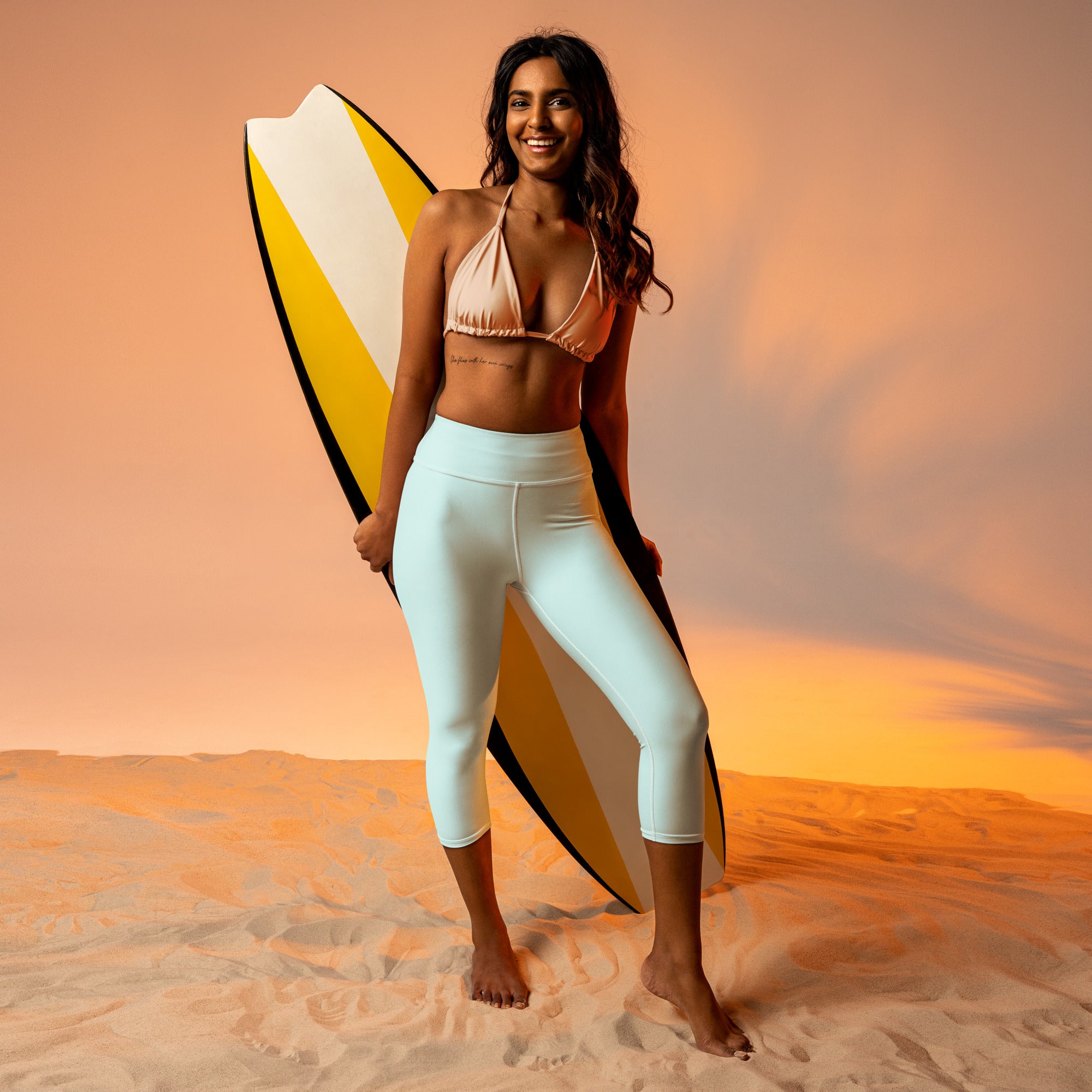A smiling woman in beach attire holds a vibrant yellow and white surfboard wearing women’s mint buttery soft yoga capri leggings