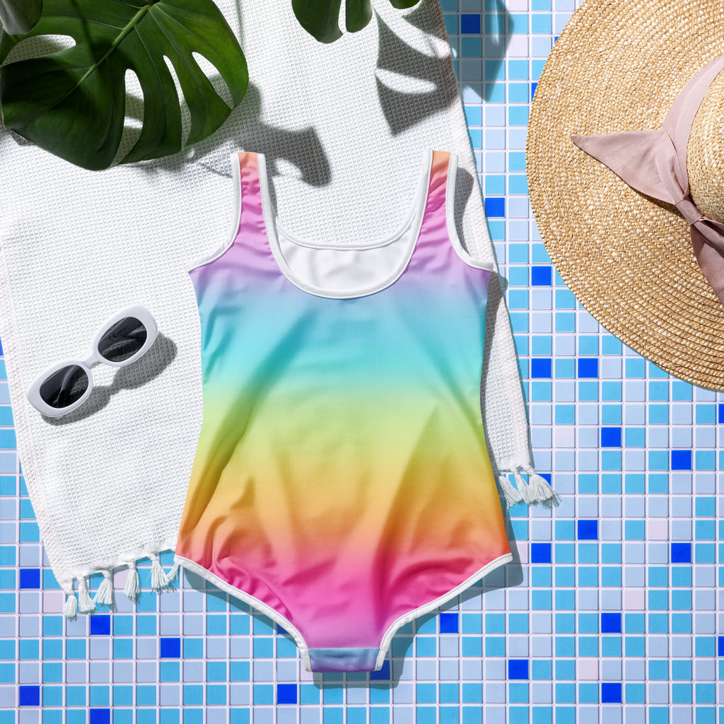 Girls on cloud 9 vibrant rainbow gradient one-piece swimsuit with white trim and scoop neckline laid on tiled surface