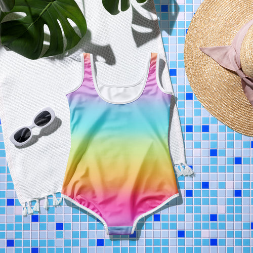 Girls on cloud 9 vibrant rainbow gradient one-piece swimsuit with white trim and scoop neckline laid on tiled surface