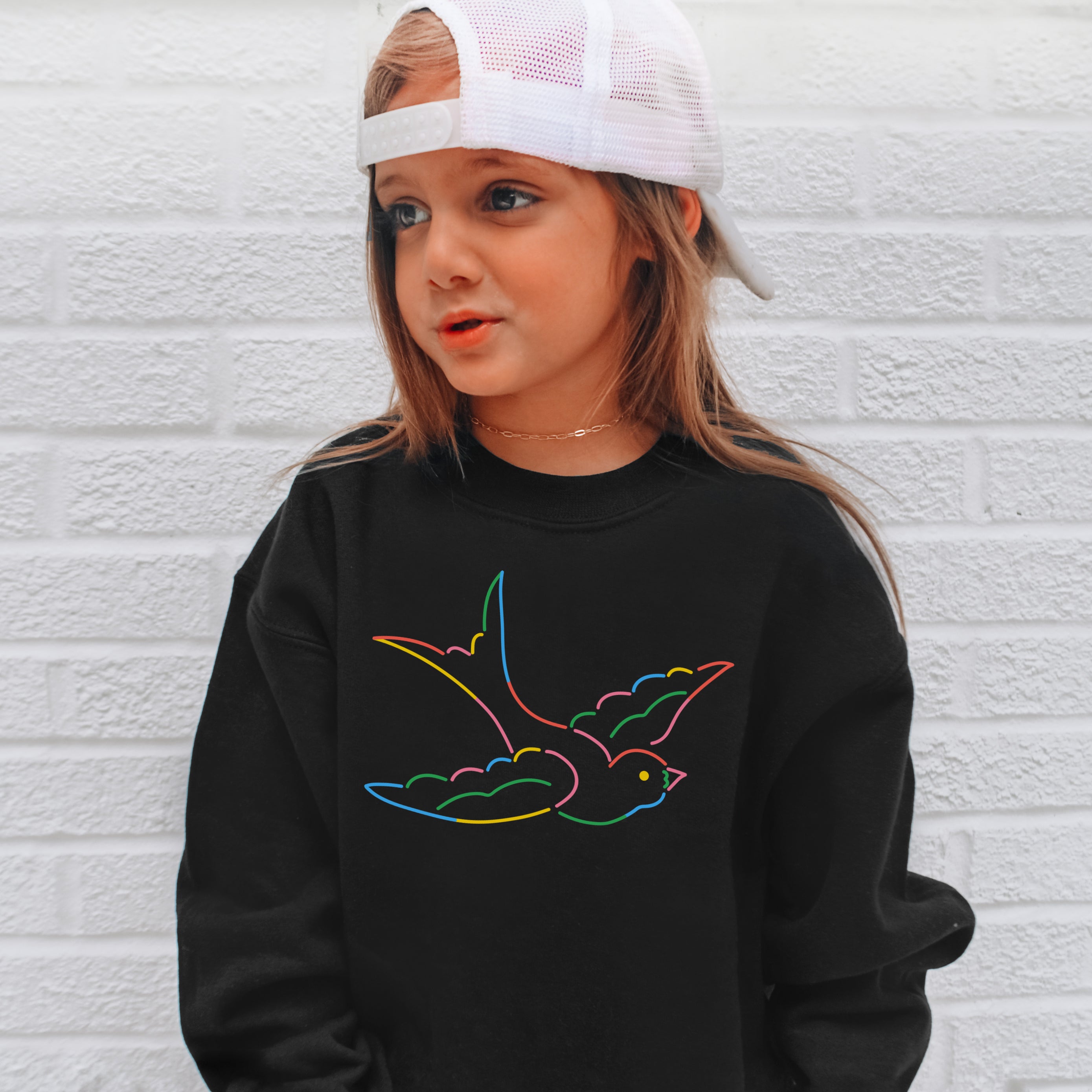 Child wearing a black sweatshirt with a colorful bird design against a white brick wall.