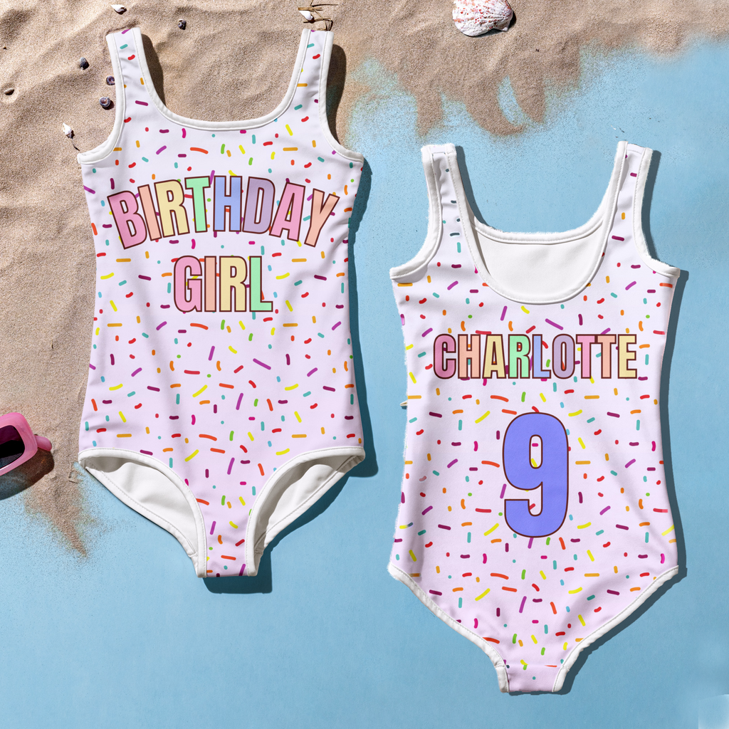 Two children's swimsuits with personalized text on a sandy and blue surface