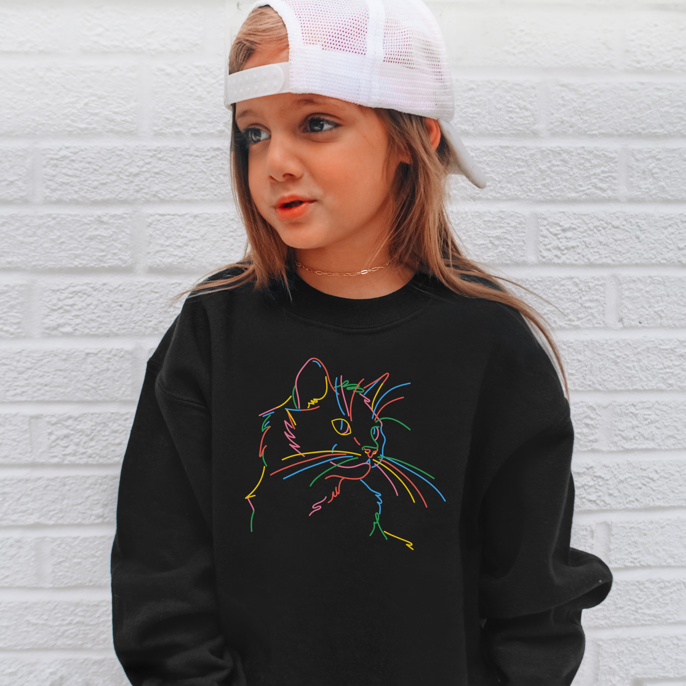 Child wearing a black sweatshirt with a colorful cat design against a white brick wall.