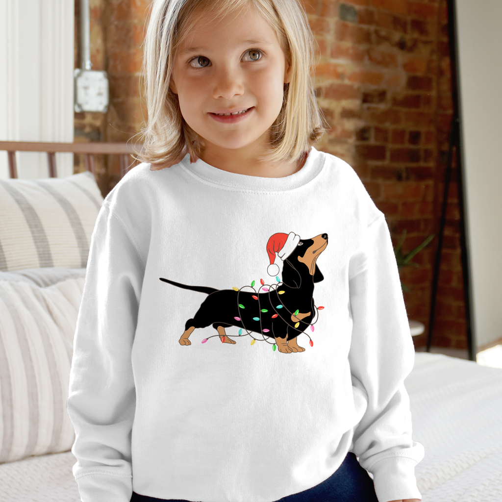 White long-sleeved kids sweatshirt with black and tan dachshund wearing a red santa hat and colorful christmas lights