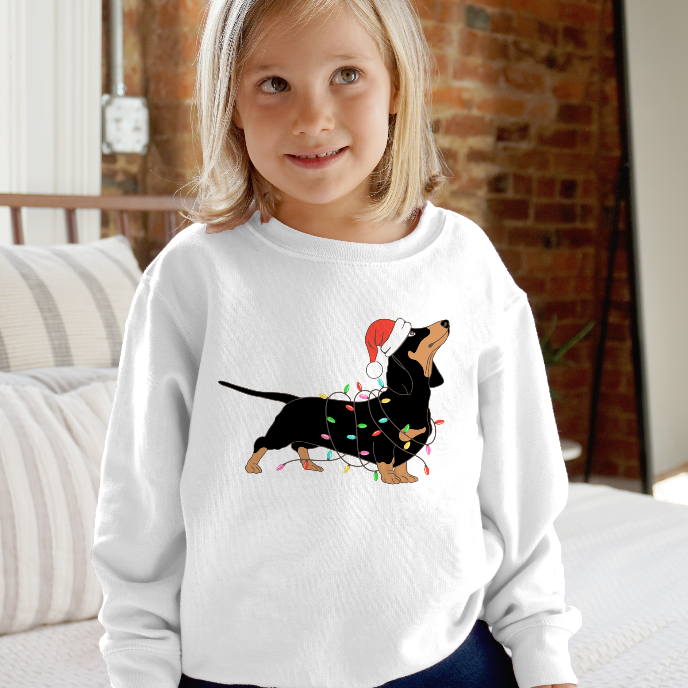 White long-sleeved kids sweatshirt with black and tan dachshund wearing a red santa hat and colorful christmas lights