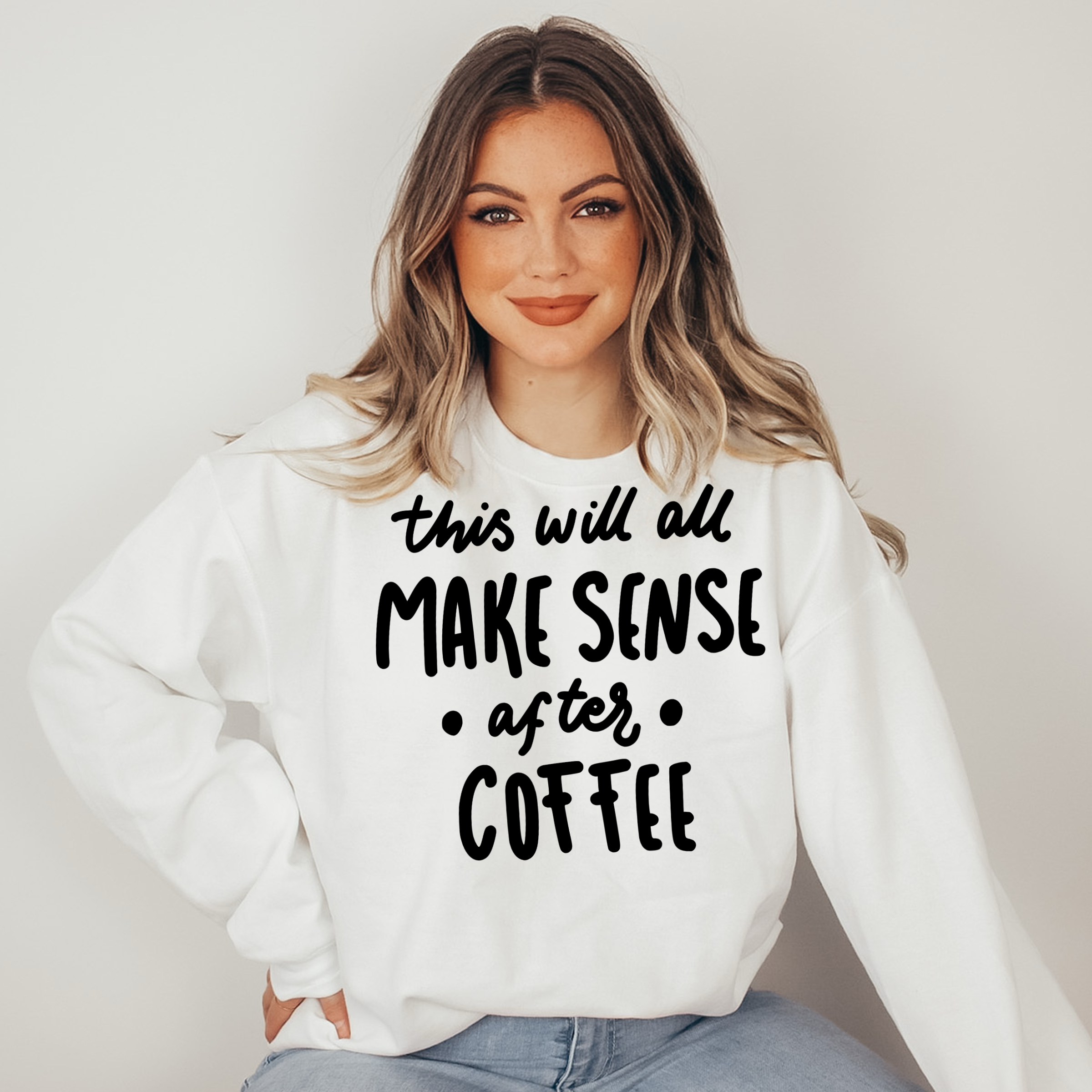 Woman wearing a white sweatshirt with text 'this will all make sense after coffee' on a plain background