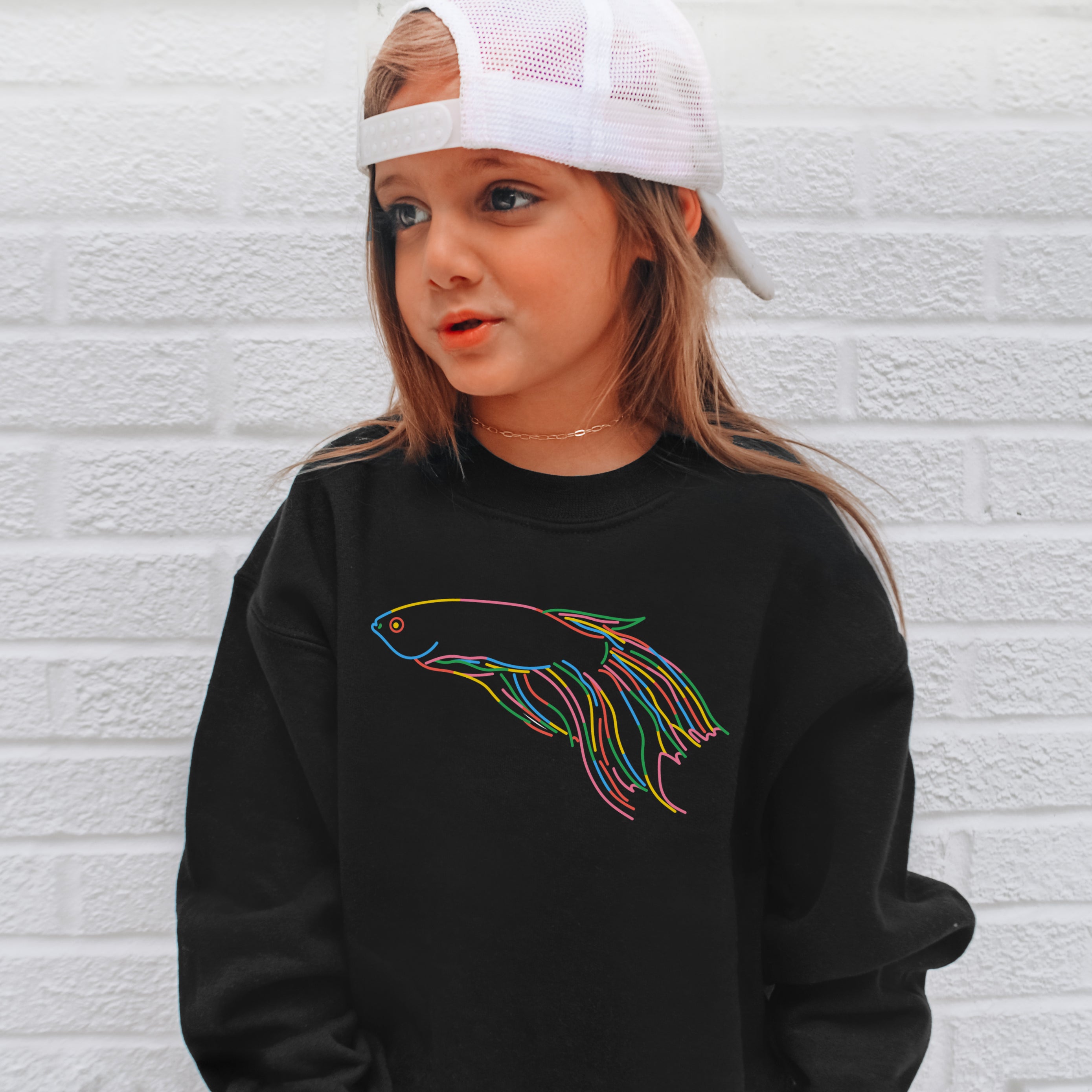 Child wearing a black sweatshirt with a colorful fish design against a white brick wall.