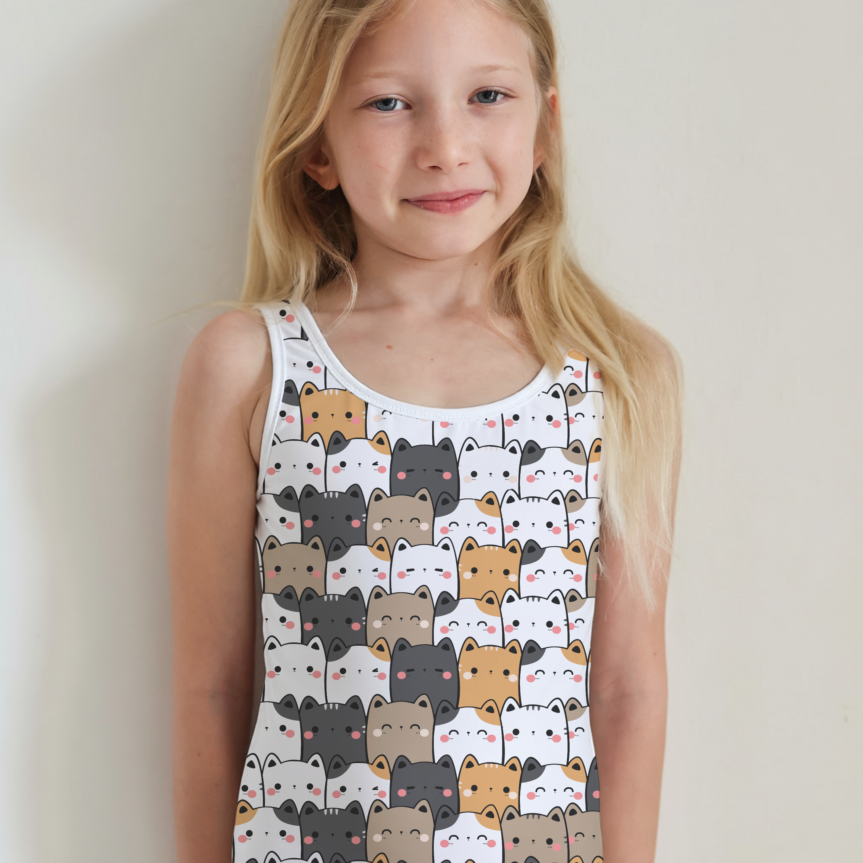 A young girl with long blonde hair stands against a plain light wall wearing a white one-piece swimsuit covered in a repeating pattern of cartoon cats in beige, brown, and gray.