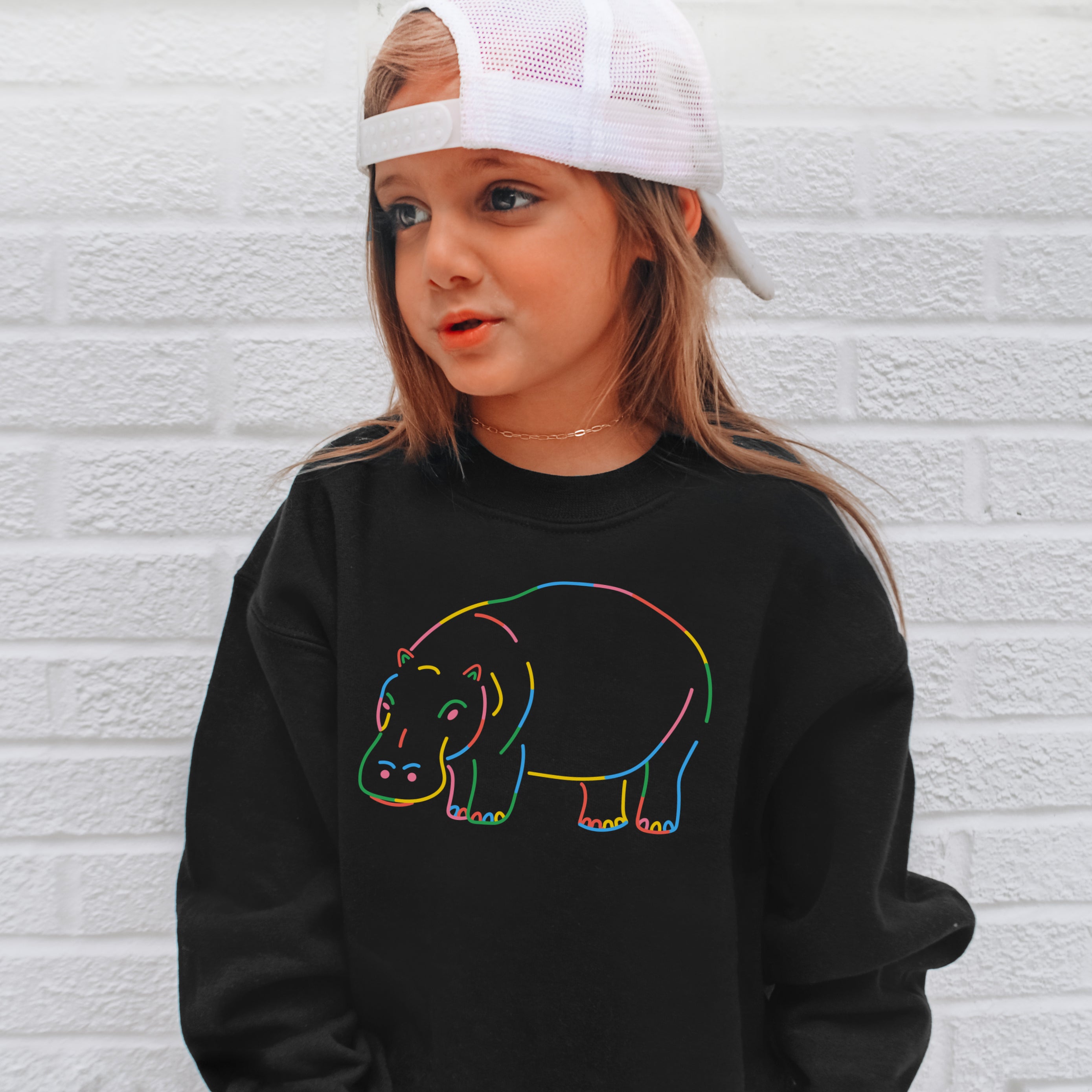 Child wearing a black sweatshirt with a colorful hippo design against a white brick wall.