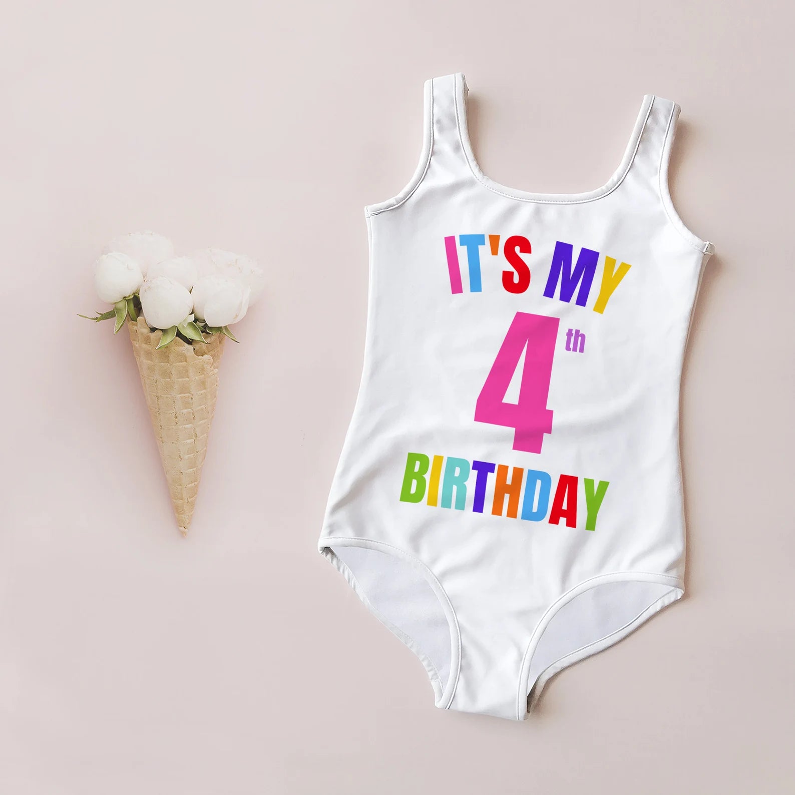 A white swimsuit for girls with 'IT'S MY 4th BIRTHDAY' printed in colorful letters on the front, displayed next to a ice cream cone with white flowers on top.