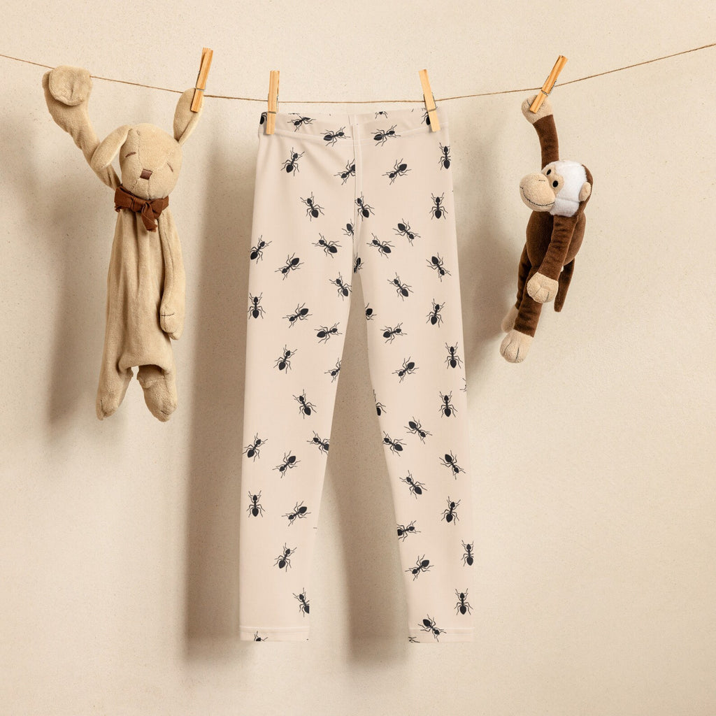 Cream-colored kids buttery soft leggings with playful black ant pattern and upf 50 plus hanging between plush toys