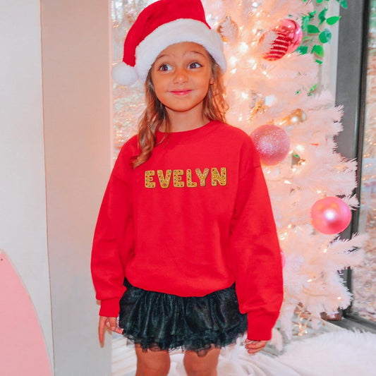 Child wearing a red sweatshirt with custom name and a Santa hat in a festive room.
