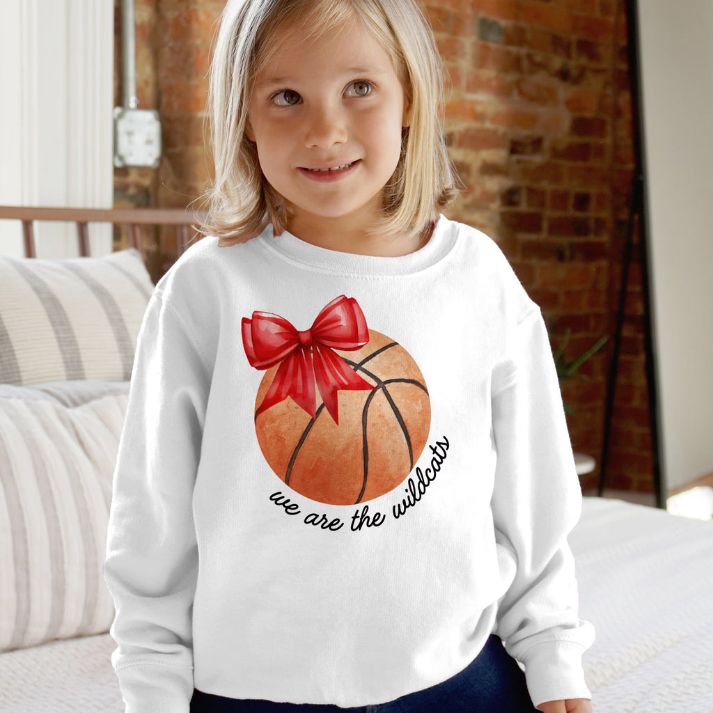 White long-sleeved sweatshirt featuring an orange basketball with a red bow and we are the wildcats text in black cursive