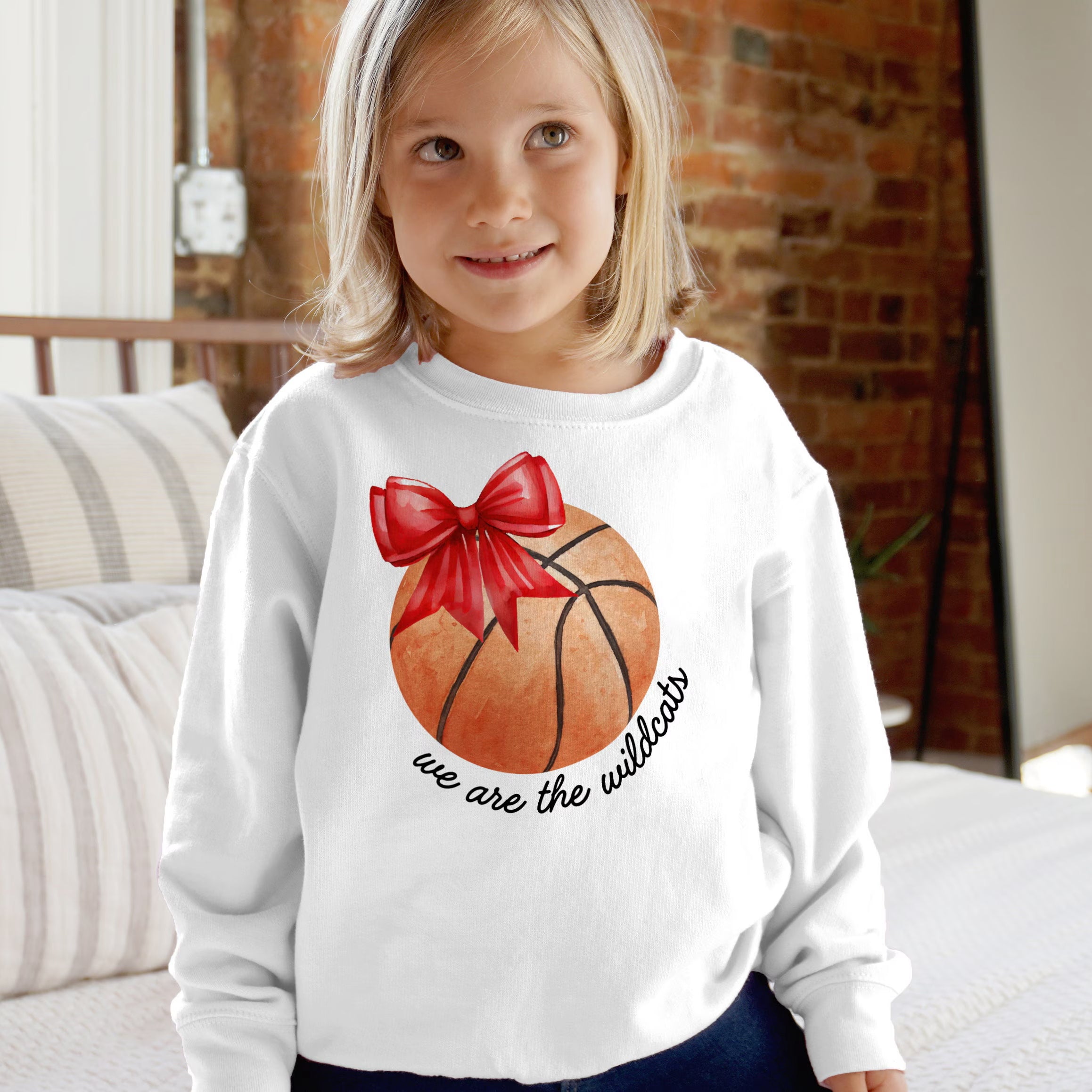 White long-sleeved sweatshirt featuring an orange basketball with a red bow and we are the wildcats text in black cursive